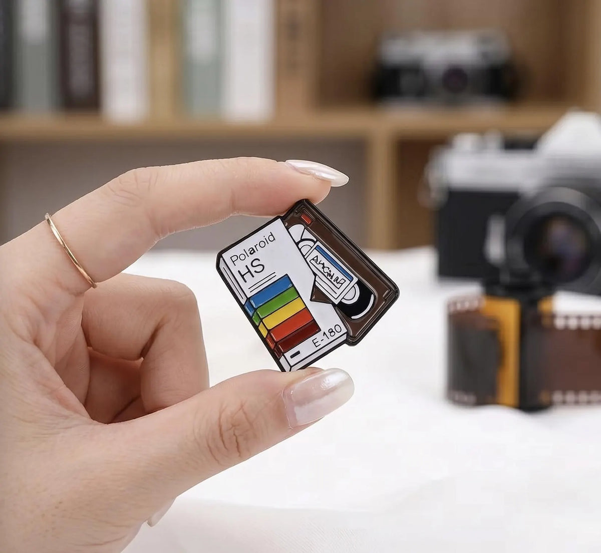 A hand holding a miniature Polaroid H5 camera memory card, with a blurred vintage camera and film strip in the background. Retro VHS Tape Enamel Pin | Vintage VCR Video Cassette Badge | 80s 90s Nostalgia Movie Lover Gift | Aesthetic Rainbow Movie Pins, Film Lover