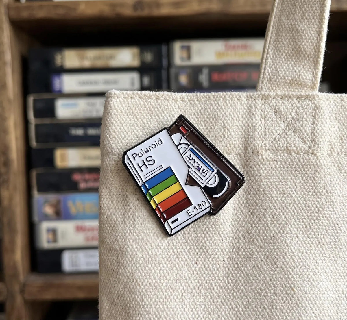 A hand holding a miniature Polaroid H5 camera memory card, with a blurred vintage camera and film strip in the background. Retro VHS Tape Enamel Pin | Vintage VCR Video Cassette Badge | 80s 90s Nostalgia Movie Lover Gift | Aesthetic Rainbow Movie Pins, Film Lover