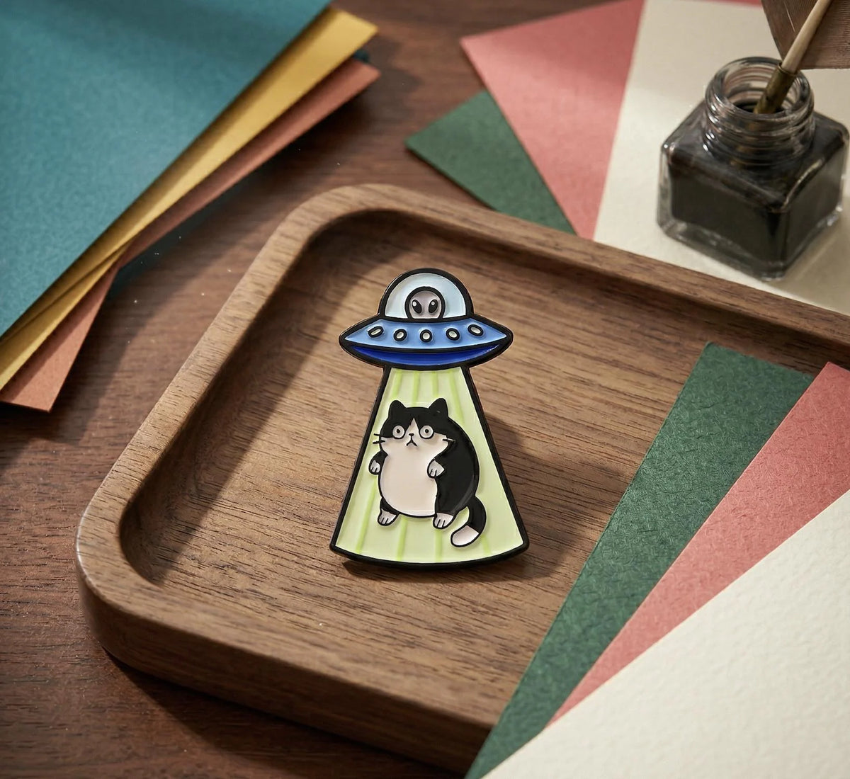 A small enamel pin shaped like a black-and-white cat under a blue UFO with green rays, displayed on a wooden tray with colorful paper and an ink bottle in the background.Black Cat Alien Enamel Pin - Space Core UFO Cat Pin for Backpacks - Cute Galaxy Kitten Pin - Unique Spooky Gift for Cat Lovers, Cosmic Cat