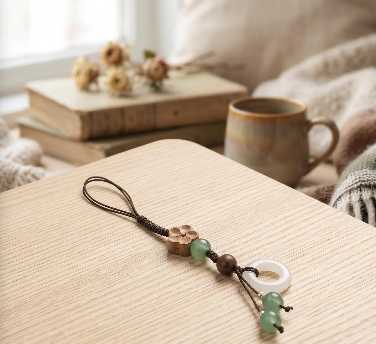 A close-up of a hand holding a black smartphone, showing a decorative phone strap with a braided cord, bronze flower charm, green and brown beads, a white jade disc, and small green beads, set against a softly blurred Japanese-style interior background. Plum Blossom Phone Charm, Wooden Flower Phone Strap, Jade Green Glass Ring Accessory, Cute Aesthetic Bag Flair, Cozy Handmade Gift for Her