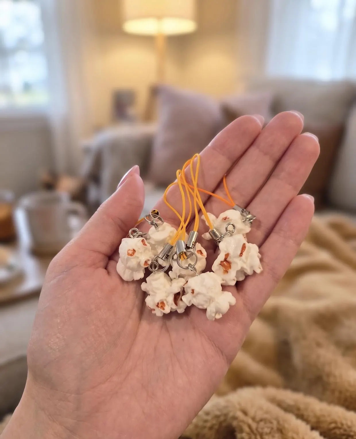 A hand holding a small cream-colored plush charm on a yellow cord, with a cozy indoor background featuring a lamp, books, and a knitted blanket. Popcorn Lover Phone Charm, Cute Miniature Food Keychain, Movie Lover Gift, Funky Cute Keychain, Corn gift, Quirky Movie Theater, Movie Night