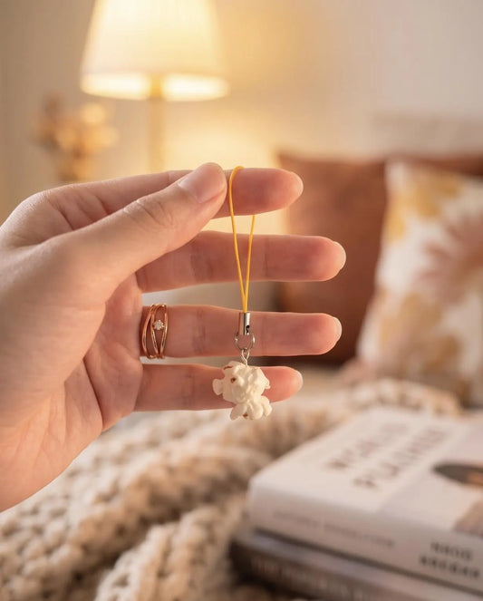 A hand holding a small cream-colored plush charm on a yellow cord, with a cozy indoor background featuring a lamp, books, and a knitted blanket. Popcorn Lover Phone Charm, Cute Miniature Food Keychain, Movie Lover Gift, Funky Cute Keychain, Corn gift, Quirky Movie Theater, Movie Night