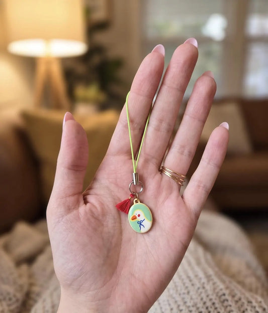 A hand holding a small oval charm with a yellow cord and tassel, featuring a cartoon bird design, against a softly blurred indoor background. Collectable Vintage Duck Charm in Bowtie, Delicate Oval Pendant, Retro Cottagecore Keychain Accessory, DIY Fashion, Duck Lovers Small Gift