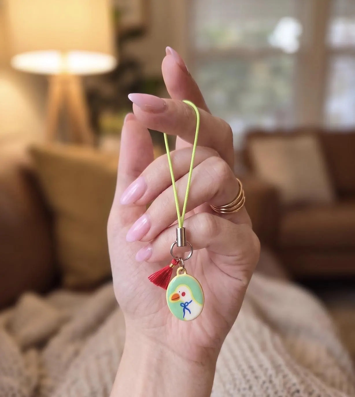 A hand holding a small oval charm with a yellow cord and tassel, featuring a cartoon bird design, against a softly blurred indoor background. Collectable Vintage Duck Charm in Bowtie, Delicate Oval Pendant, Retro Cottagecore Keychain Accessory, DIY Fashion, Duck Lovers Small Gift