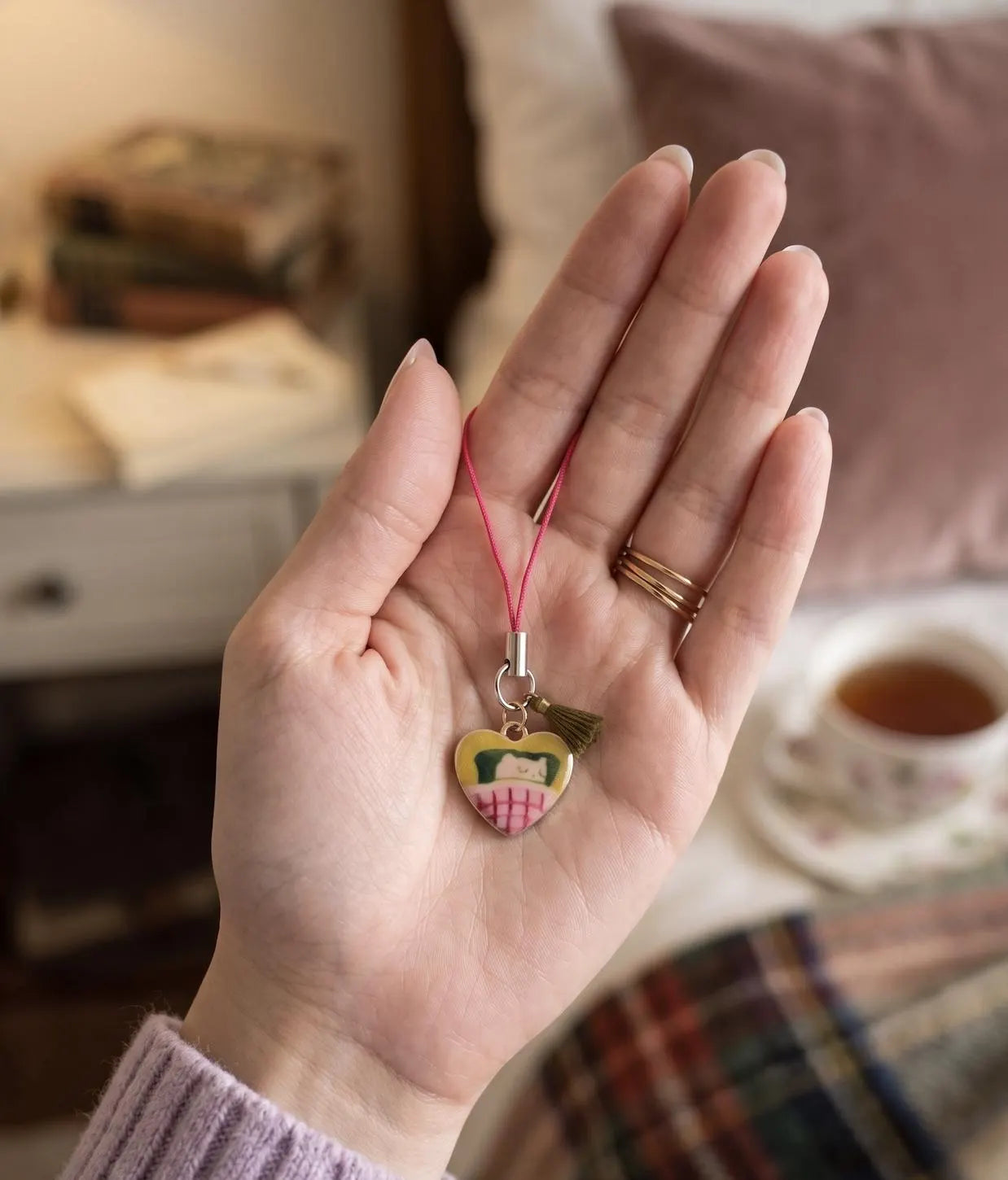 A hand with pink manicured nails holds a small heart-shaped charm with a pink strap. The charm features a yellow face with black hair and a pink-and-white striped skirt, set against a softly blurred bedroom background. Professional Cozy Napper Cat Phone Charm, Nap Lovers Gift, Vintage pendants and Charms, Jewellery Accessory, Introvert Keychain, Lazy Vibes