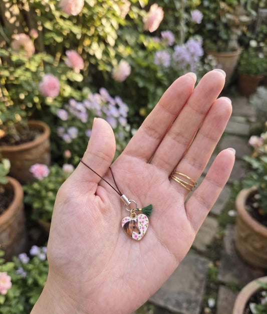 A hand holding a small heart-shaped charm with a floral pattern and green tassel on a black cord, with a blurred garden background. Antique Style Equestrian Charm, Vintage Botanical Horse Keychain, Eclectic Floral Pony Accessory, Horse Lover, Nostalgic Enamel Phone Strap