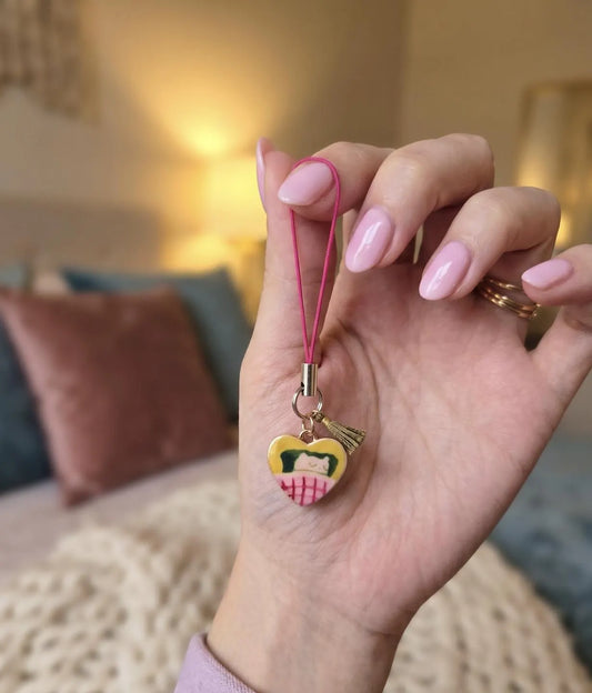A hand with pink manicured nails holds a small heart-shaped charm with a pink strap. The charm features a yellow face with black hair and a pink-and-white striped skirt, set against a softly blurred bedroom background. Professional Cozy Napper Cat Phone Charm, Nap Lovers Gift, Vintage pendants and Charms, Jewellery Accessory, Introvert Keychain, Lazy Vibes