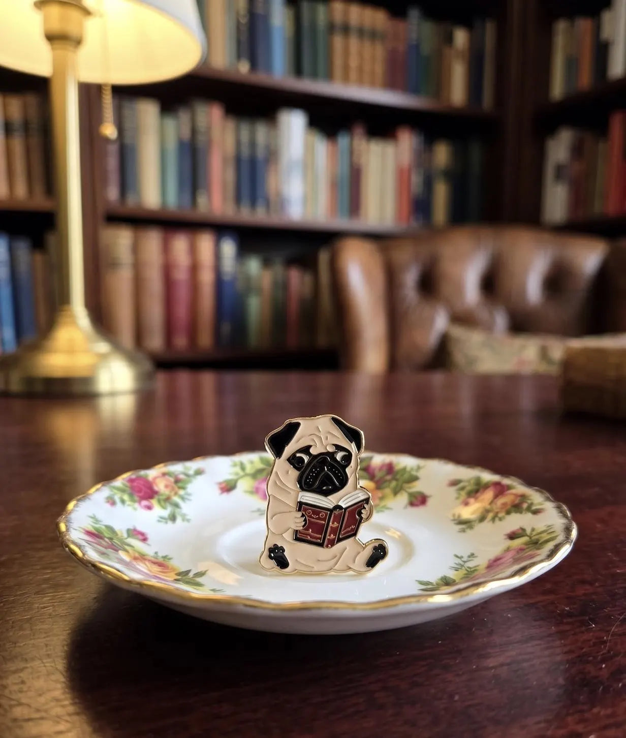 A small enamel pin shaped like a pug dog reading a book, displayed on a floral porcelain saucer on a wooden table with a blurred library background. Pugs & Prose: Cute Reading Pug Enamel Pin | Bookworm Dog Lapel Badge for Book Lovers, Reading Smart Dog, Tote Accessories, Gift for Friends