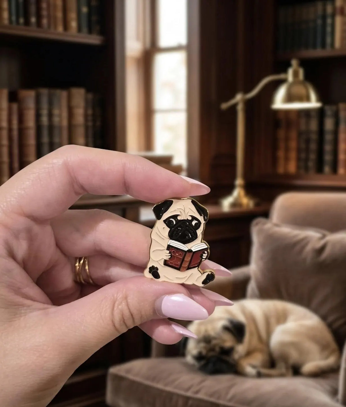 A small enamel pin shaped like a pug dog reading a book, displayed on a floral porcelain saucer on a wooden table with a blurred library background. Pugs & Prose: Cute Reading Pug Enamel Pin | Bookworm Dog Lapel Badge for Book Lovers, Reading Smart Dog, Tote Accessories, Gift for Friends