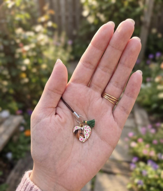 A hand holding a small heart-shaped charm with a floral pattern and green tassel on a black cord, with a blurred garden background. Antique Style Equestrian Charm, Vintage Botanical Horse Keychain, Eclectic Floral Pony Accessory, Horse Lover, Nostalgic Enamel Phone Strap