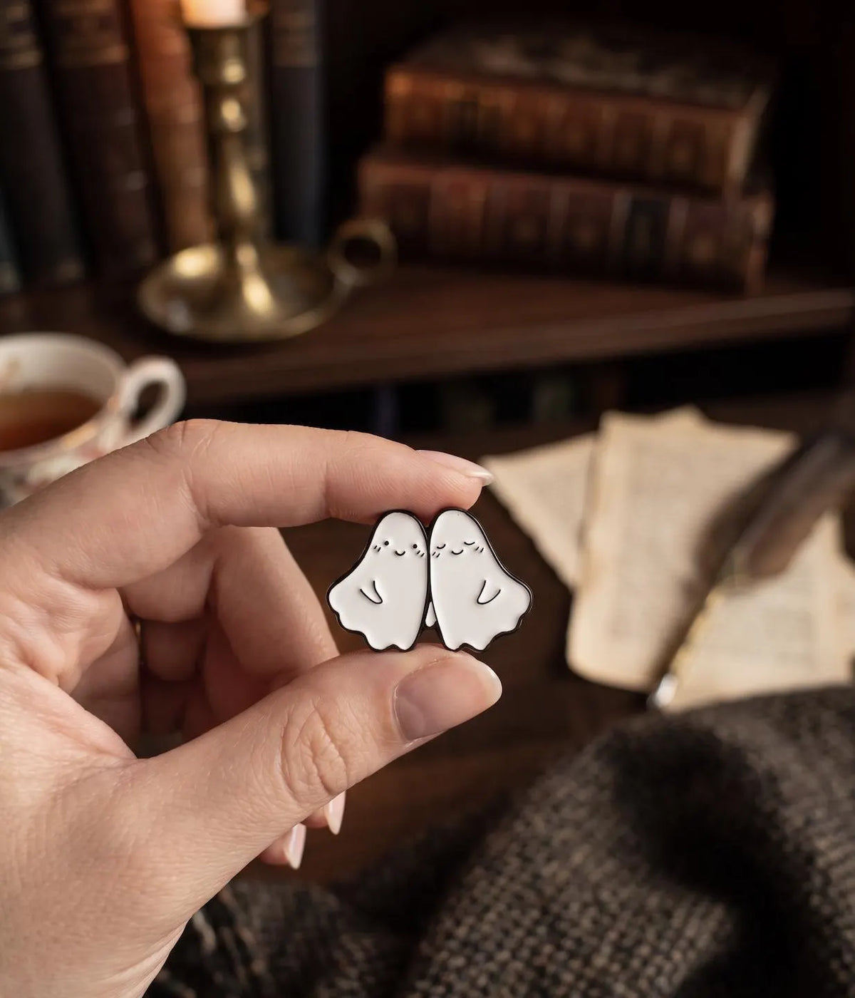 A hand holds a pair of small white ghost-shaped enamel pins with simple black line drawings, set against a cozy, blurred background of books, a cup of tea, and a feather quill. Forever Ghost Besties Enamel Pin | Soft Goth Aesthetic Halloween Pin | RIP Ghosts Spooky Lovers Gift | Cute Matching Best Friend Lapel Pin best friend pin, matching bff pins, cute ghost pin, halloween pin, soft goth pin, friendship gift, bestie gift, spooky cute gift, backpack pin, matching pins, gift for bff, hard enamel pin, kawaii