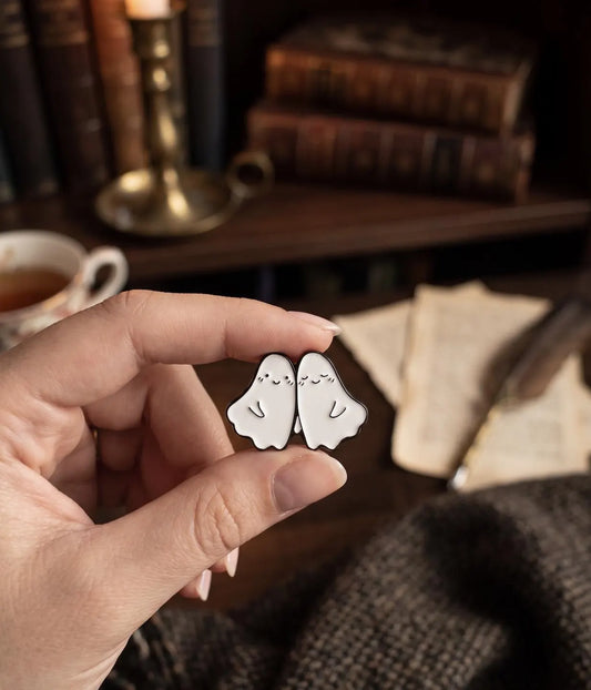 A hand holds a pair of small white ghost-shaped enamel pins with simple black line drawings, set against a cozy, blurred background of books, a cup of tea, and a feather quill. Forever Ghost Besties Enamel Pin | Soft Goth Aesthetic Halloween Pin | RIP Ghosts Spooky Lovers Gift | Cute Matching Best Friend Lapel Pin best friend pin, matching bff pins, cute ghost pin, halloween pin, soft goth pin, friendship gift, bestie gift, spooky cute gift, backpack pin, matching pins, gift for bff, hard enamel pin, kawaii