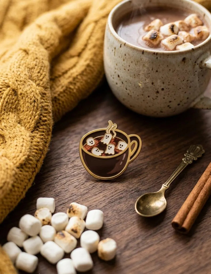 A hand with pink manicured nails holds a small enamel pin shaped like a cup of hot chocolate with marshmallows, set against a cozy background of a coffee mug, knit blanket, and wooden table. GhostMallow Hot Chocolate Enamel Pin | Coffee Lover Gift, Halloween Pin | Cute Spooky Lovers Gift | Ghostly Bag Accessories, Marshmallows